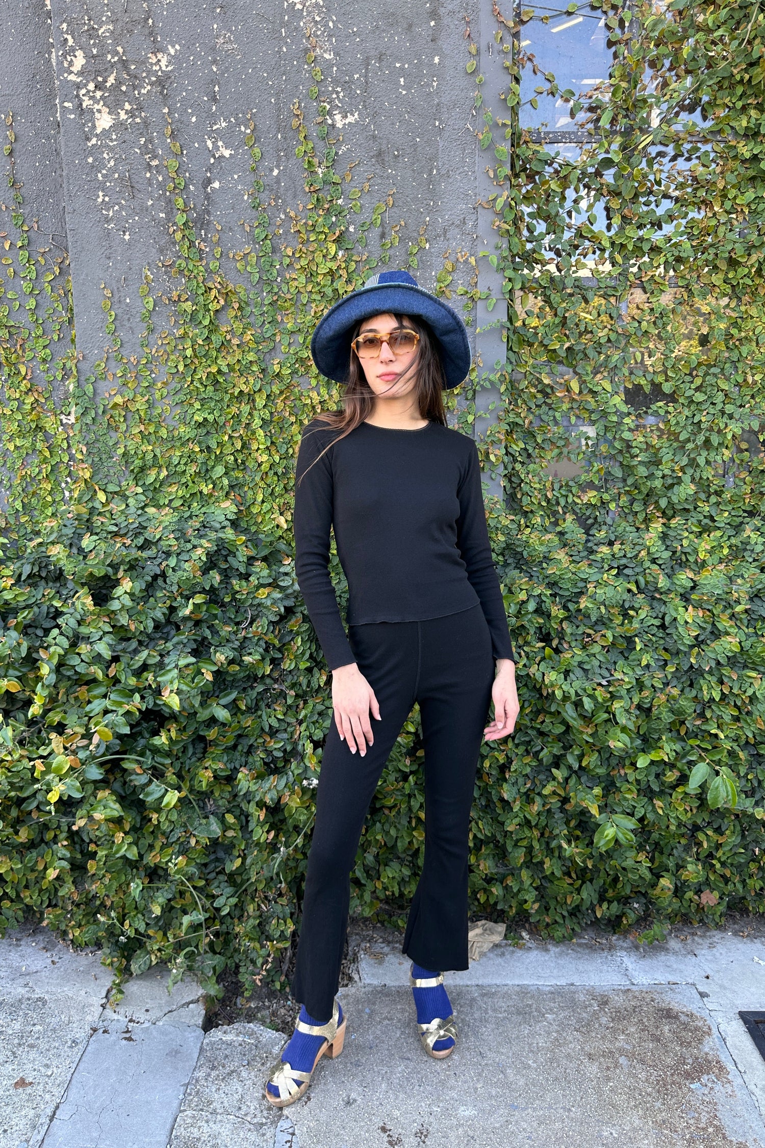 Person wearing a black outfit and blue hat standing on a sidewalk with green bushes in the background
